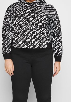 Czarna i biała bluza z kapturem Calvin Klein Jeans z nadrukiem całopowierzchniowym logo, noszona z prostymi czarnymi spodniami.
