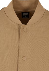 Veste en coton couleur fauve avec boutons-pression et col côtelé, affichant l'étiquette de marque Urban Classics et l’étiquette taille medium à l’intérieur du col.