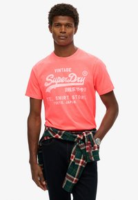 T-shirt corail vif avec le logo vintage "SuperDry" en blanc. Manches courtes, col rond. Chemise en flanelle à carreaux nouée autour de la taille avec des tons de bleu et de vert.