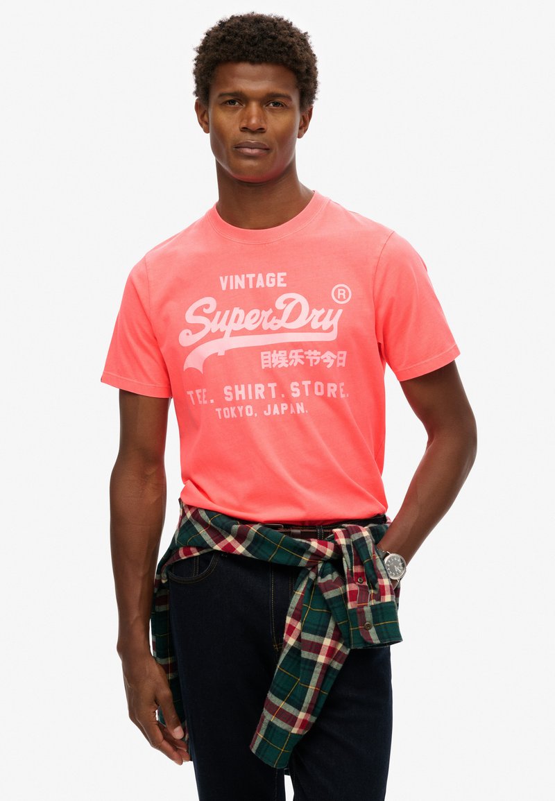 T-shirt corail vif avec le logo vintage "SuperDry" en blanc. Manches courtes, col rond. Chemise en flanelle à carreaux nouée autour de la taille avec des tons de bleu et de vert.