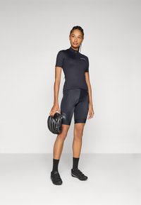 Schwarzes Radtrikot mit kurzen Ärmeln und einem Reißverschluss auf der Vorderseite, kombiniert mit schwarzen gepolsterten Shorts und einem schwarzen Helm in der Hand. Radschuhe vervollständigen das Outfit.