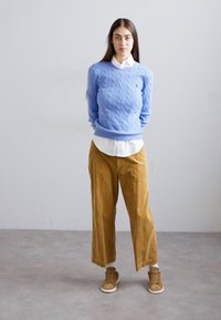 Moteris stovi su rankomis už nugaros, apsirengusi mėlynos spalvos megztiniu su rašteliu, baltu marškinėliu, geltonais plačiais keliais ir rudiems sportbačiais.