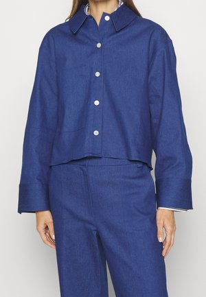 Femme portant une veste-chemise boutonnée bleue assortie et un pantalon taille haute avec des boutons argentés, debout devant un fond blanc uni.