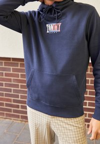 Navy blue pullover hoodie with drawstring neck, featuring a white and red "TOMMY JEANS" logo. Includes a front kangaroo pocket and ribbed cuffs.