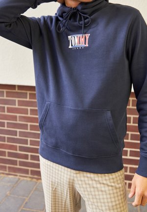Haut à capuche bleu marine avec cordon de serrage, arborant un logo "TOMMY JEANS" en blanc et rouge. Comprend une poche kangourou à l'avant et des poignets côtelés.