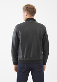 Chaqueta de cuero negra con un cuello y puños acanalados, que presenta una textura suave y un diseño ajustado, llevada con jeans oscuros.