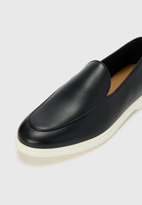 Fekete bőr, slip-on cipő sima textúrával, kerek orral és kontrasztos fehér gumitalppal. Minimalista dizájn, látható díszítések nélkül.