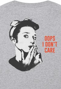 T-shirt grigia con una grafica in bianco e nero di una persona con fascia per capelli, mano sul viso, e il testo "OOPS NON ME NE FREGA" in arancione brillante.