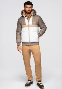 Padded jacket w kolorach brązowym, białym i kremowym, z zapięciem na zamek, kapturem oraz jednolitymi blokami kolorów; zestawiona z beżowymi spodniami i ciemnymi sneakersami.