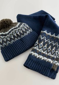 Tamsiai mėlynas megztas kepurė su rude pom-pom ir geometriniais baltais raštais; derančios tamsiai mėlyną šalikas su panašiais motyvais ir įribiuotais kraštais.