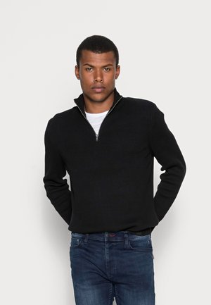 Svart ribbestrikket genser med glidelås og strukturert stoff, kombinert med blå jeans. Designet har høy krage og en tettsittende silhuett.