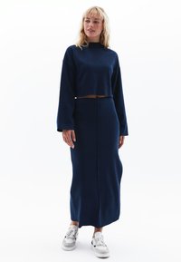 Navyblauwe cropped trui met lange mouwen en een hoge halslijn, gecombineerd met een vloerlange rok met een voornaaddetail. Witte sneakers.