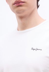 Κλείσιμο κοντινού πλάνου ενός α τόμου που φοράει λευκό μπλουζάκι με τη λέξη "Pepe Jeans" κεντημένη με μαύρα γράμματα στο στήθος.