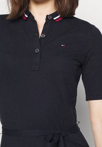 Navy Polohemd mit einem gestreiften Kragen in Rot, Weiß und Blau, ausgestattet mit einer Knopfleiste mit vier Knöpfen und einem gebundenen Taillendetail.