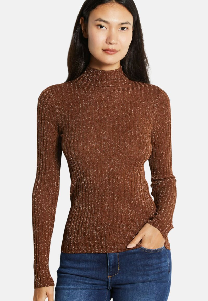 Vrouw met lang donker haar, gekleed in een aangesloten bruine geribbelde coltrui met metalen draadjes en een blauwe spijkerbroek, met één hand in haar broekzak.