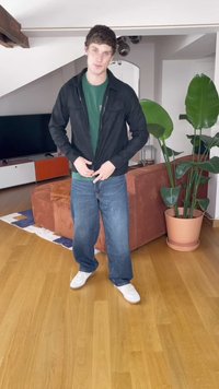 Fekete cipzáras dzseki zöld ing fölött, kék farmernadrággal és fehér tornacipővel. A háttérben barna kanapé és nagy cserepes növények láthatók.