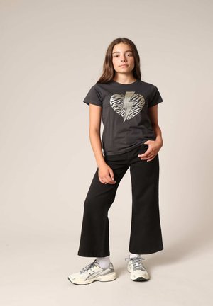 Grijze t-shirt met een metallic gouden hart en bliksemschicht ontwerp, gecombineerd met zwarte wijde broek en grijze sportschoenen.