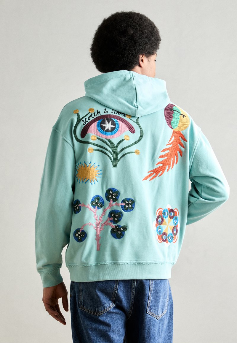 Persona che indossa un hoodie azzurro chiaro con disegni ricamati colorati, tra cui un occhio, piante e forme abstracte sul retro.