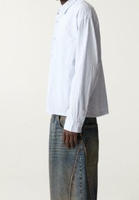 Jasnoniebieska, pomarszczona bawełniana koszula zapinana na guziki o luźnym kroju, z kieszonką na piersi i długimi rękawami. Noszona z podartych dżinsów.