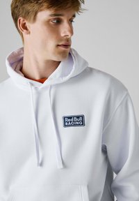 Felpa bianca con cappuccio e tasca a kangaroo; presenta un logo ricamato blu "Red Bull Racing" sul petto. Tessuto morbido, vestibilità casual.