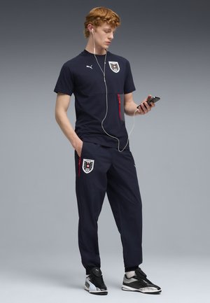 Jonge man die zwarte Puma sportkleding draagt met logo's van de Oostenrijkse voetbalbond, kijkt naar zijn telefoon met bedrade oordopjes, staand tegen een grijze achtergrond.