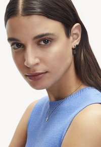 Le modèle porte des boucles d'oreilles noires avec des motifs floraux et circulaires. Elle a les cheveux bruns raides et est vêtue d'un haut sans manches à rayures bleu.