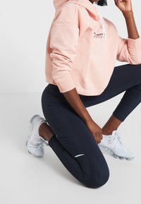 Jasnoróżowa krótka bluza z kapturem z białymi sznurkami i logo. Czarne legginsy o smukłej linii. Białe sneakersy z teksturowanymi detalami.