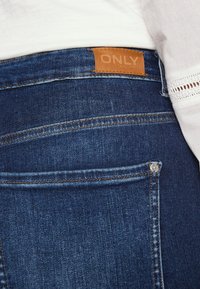 Jeans de ganga azul escuro com uma etiqueta de couro castanha rotulada "ONLY". Costuras visíveis e um bolso traseiro com um detalhe de botão prateado.