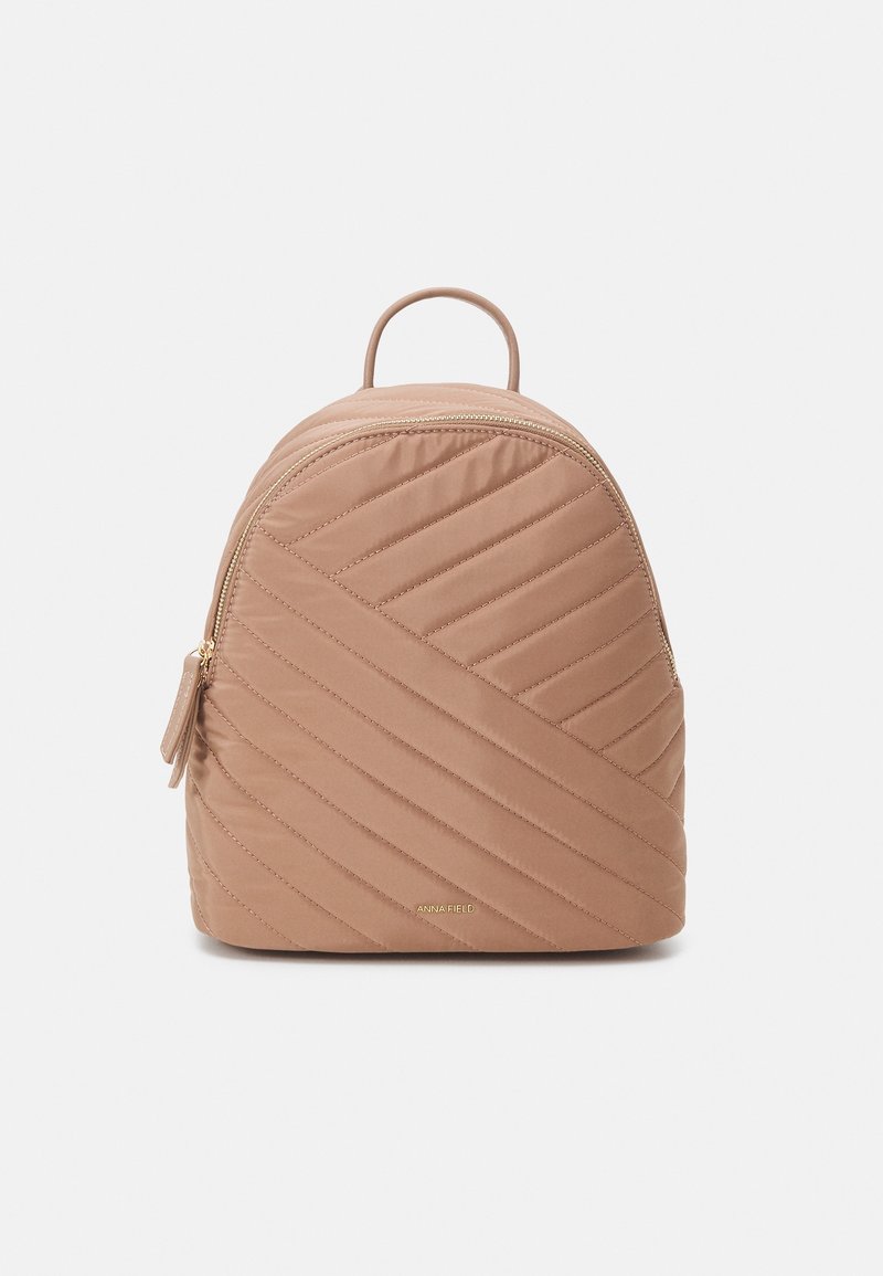 Lite beige kviltad ryggsäck med diagonal sömnad, guldfärgad dragkedja, handtag upptill och diskret guld logotyp på framsidan.