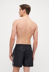 Pantaloni da bagno neri con vita elasticizzata, tasche laterali e strisce laterali a contrasto. Il tessuto è morbido e leggero.