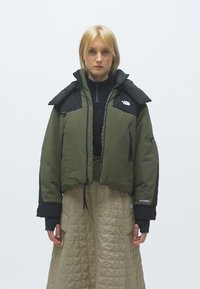 Casaco acolchoado verde-oliva com acabamentos pretos, gola alta, fecho de correr na frente e bolsos laterais. Combinado com uma saia longa de tom beige texturizada.