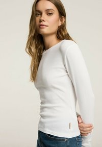 Biała, długo rękawowa bluzka z prążkowanej tkaniny, z okrągłym dekoltem i małą metką z boku, zestawiona z niebieskimi dżinsami.