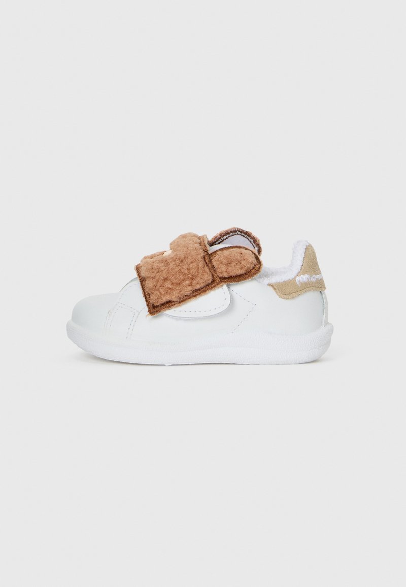 Baskets blanches avec une sangle en velcro brun doux, bout arrondi, dessus texturé et accents en polaire au niveau du col. Semelle en caoutchouc pour une meilleure adhérence.