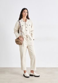 Jumpsuit color crema con chiusura a bottoni, due tasche sul petto e vita legata. Mocassini neri con tacco e borsa a tracolla marrone con un motivo.