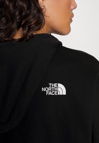 Sudadera negra de tejido suave, con un logo blanco de The North Face en la parte posterior cerca del cuello. Diseño con capucha y costuras visibles.
