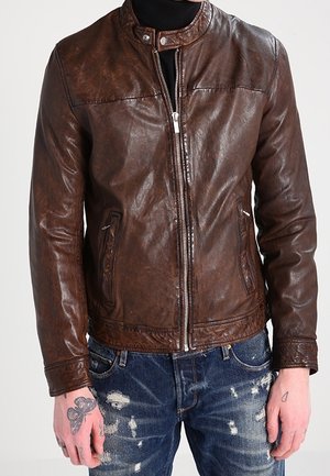 Mand iført en brun læderjakke med lynlåslukning, kombineret med slidte blå jeans og synlige tatoveringer på hænderne.