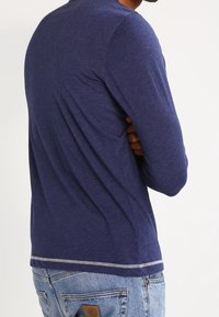 Dlouhý rukáv námořnický modrý košile vyrobená z měkké tkaniny, s kulatým výstřihem a kontrastním prošitím na lemu. Nosená s džínovými kalhotami.