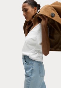 T-shirt bianco a collo a V abbinato a jeans di denim blu chiaro, con un cappotto marrone con bottoni neri appoggiato sulla spalla. Design minimalista.