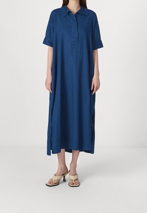 Femme portant une robe bleue ample mi-mollet avec manches courtes et col, associée à des sandales à talons beiges.