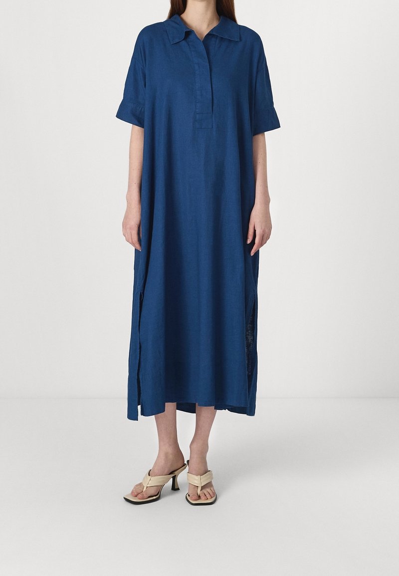 Femme portant une robe bleue ample mi-mollet avec manches courtes et col, associée à des sandales à talons beiges.