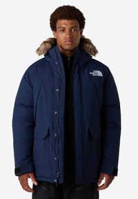 Giacca isolante blu navy con cappuccio foderato in pelliccia sintetica, bottoni a pressione, due tasche frontali e logo sul lato sinistro del petto. Materiale resistente e impermeabile.