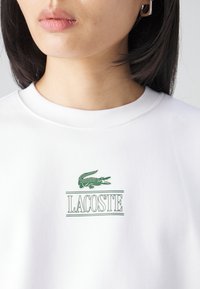 Fehér környakú pulóver, zöld hímzett Lacoste logóval és krokodil mintával. Simaságot és kényelmet biztosító pamut anyag, laza szabással.