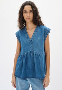 Džínová tuniková blúzka v modrej farbe s výstrihom do V, krátkymi rukávmi a peplum pásom. Má viditeľné švy a voľný strih.