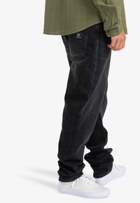 Persona vestida con jeans negros holgados, zapatillas blancas y una camiseta de manga larga verde, de pie con la mano derecha relajada a su lado.