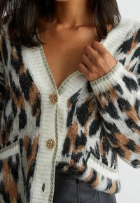 Gilet en maille douce et texturée avec un audacieux motif léopard noir, marron et crème. Des boutons dorés et des poches complètent le design.