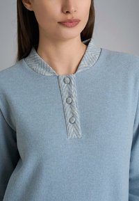 Maglione azzurro chiaro con colletto in tessuto a trama a zig-zag. Presenta tre bottoni sul davanti e un tessuto morbido simile al pile.