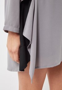 Mano descansando cerca del muslo, vistiendo un vestido gris claro con una capa interior negra y un detalle de tela fluida en capas.