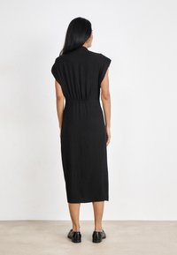 Robe noire mi-longue avec manches courtes, tissu texturé, taille cintrée et silhouette droite. Portée avec des chaussures noires.
