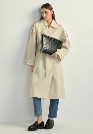 Kobieta w beżowym płaszczu trenczu, niebieskich dżinsach, czarnych mokasynach, trzymająca dużą czarną torebkę kopertówkę z fakturą na jasnym, jednolitym tle.