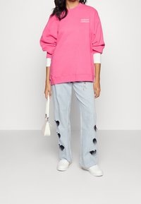 Person som bär en ljusrosa Hollister California-tröja, ljusblå jeans med hjärtformade utskärningar, vita sneakers och håller en vit väska.